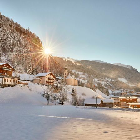 Eine friedliche Winterlandschaft mit einem Dorf in den schneebedeckten Bergen, einer Kirche, die auf einem Hügel steht, und der Sonne, die hell am Himmel scheint.