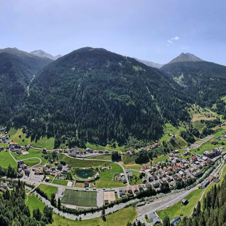 Luftaufnahme einer kleinen Stadt in den Schweizer Alpen, umgeben von dichten Wäldern und Bergen. Die Stadt verfügt über ein Fußballfeld, einen Teich und zahlreiche Gebäude.