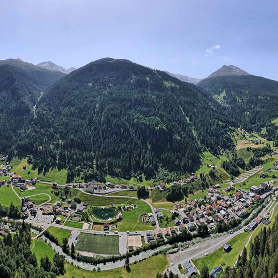 Luftaufnahme einer kleinen Stadt in den Schweizer Alpen, umgeben von dichten Wäldern und Bergen. Die Stadt verfügt über ein Fußballfeld, einen Teich und zahlreiche Gebäude.