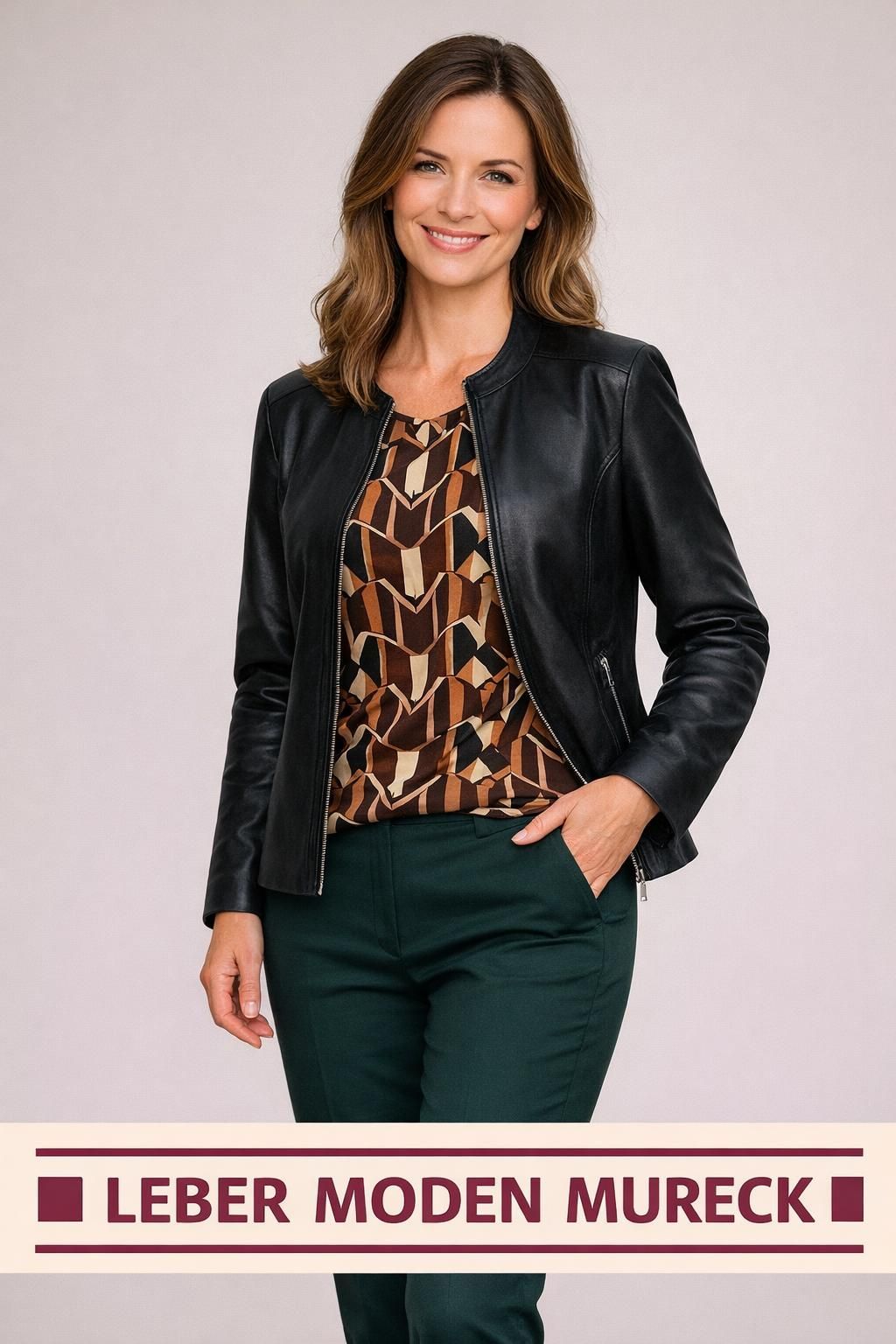 Eine Frau trägt eine schwarze Lederjacke mit Reißverschluss, gepaart mit einem gemusterten Oberteil und grüner Hose. Sie lächelt und posiert für ein Foto.