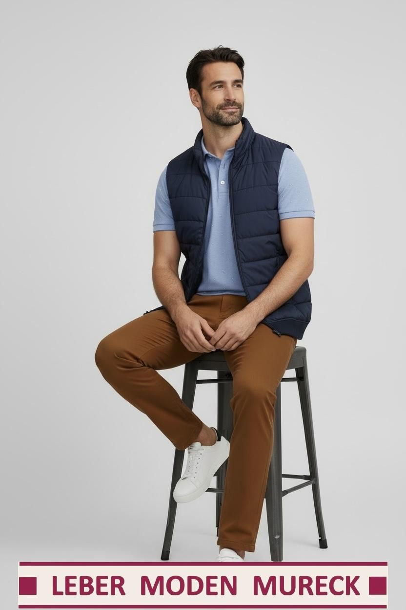 A man is sitting on a stool wearing a blue shirt, brown pants, and a navy blue vest. He is smiling slightly.
