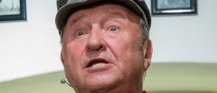 Ein Mann in einer Militäruniform mit einer grauen Mütze sieht überrascht in die Kamera.