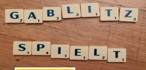 Scrabble-Steine bilden die Wörter "GABLIZ" und "SPIELT" auf einer Holzoberfläche.
