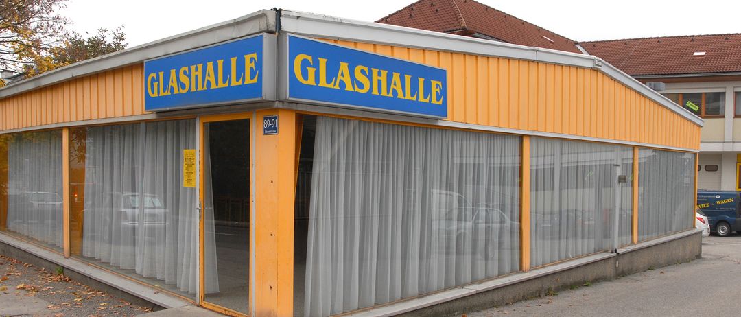 Die Vorderseite eines gelben Gebäudes hat ein Schild mit der Aufschrift 'Glashalle'. Das Gebäude hat eine Glastür, eine Glaswand und weiße Vorhänge. Die Nummer '89-91' steht auf der Glastür. Hinter dem Gebäude sind einige Autos vor einem Gebäude mit einem braunen Dach geparkt.