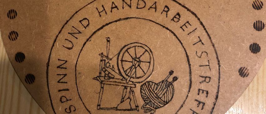 Eine Nahaufnahme eines braunen Etiketts mit einer altmodischen Spinnmaschine mit einem Rad und Garnrollen, umgeben von dem Text 'SPINNEREI UND WEBEREISTRASSE'. Das Etikett hat ein leicht abgenutztes Aussehen.