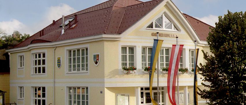 Ein gelbes Gebäude mit rotem Dach und weißer Umrandung hat mehrere Fenster, einen Fahnenmast mit Flaggen und eine Namenstafel, die 'GeindebAm' liest. Unter den Fenstern befinden sich Pflanzen in Töpfen.