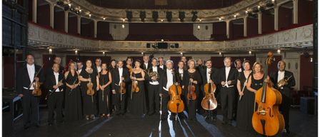 Ein großes Orchester steht in einem Halbkreis auf der Bühne und hält verschiedene Instrumente in den Händen, unter Scheinwerfern.