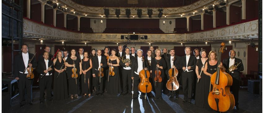 Ein großes Orchester steht in einem Halbkreis auf der Bühne und hält verschiedene Instrumente in den Händen, unter Scheinwerfern.