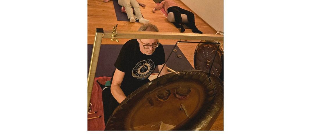Eine Gruppe von Menschen praktiziert Yoga auf einem Holzboden, angeleitet von einem Mann, der eine Glocke spielt. Der Raum hat ein großes Buddha-Poster und eine Holztür.