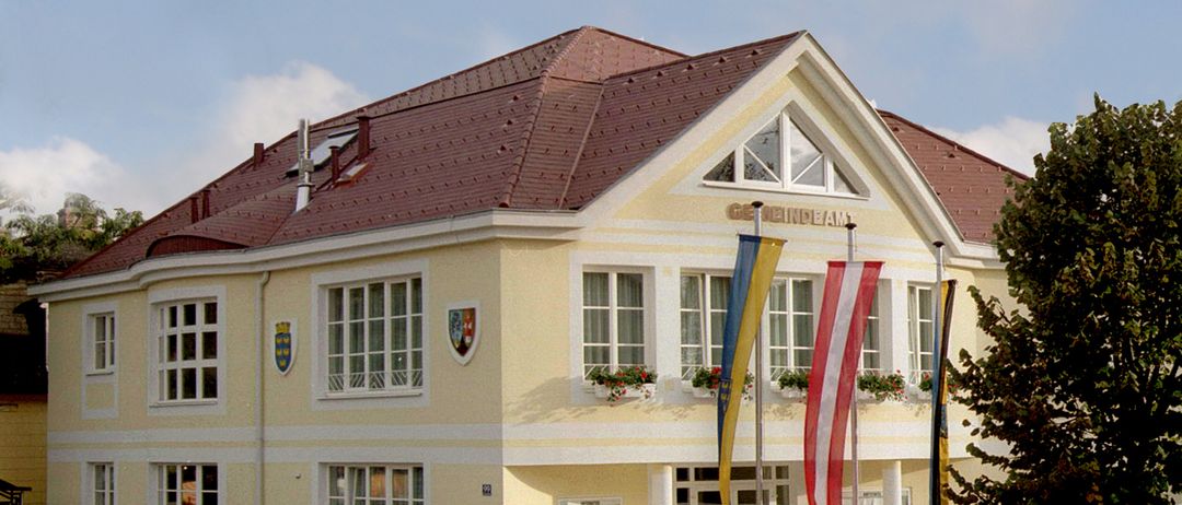 Ein gelbes Gebäude mit einem roten Dach verfügt über mehrere Fenster und dekorative Schilde über dem Eingang. Vor dem Gebäude befinden sich drei Fahnen auf Stangen, mit einem Gehweg zum Eingang.