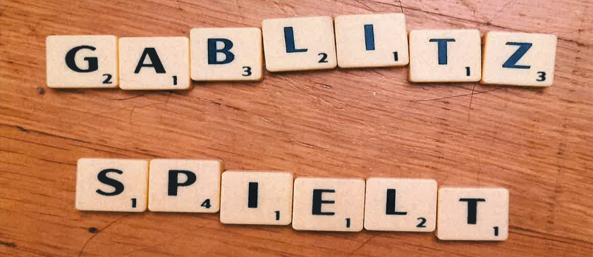 Scrabble-Steine bilden auf einer Holzoberfläche 'GABLIZT'. Darunter bilden die Steine 'SPIELT'. Unten ist ein gelber Rand.