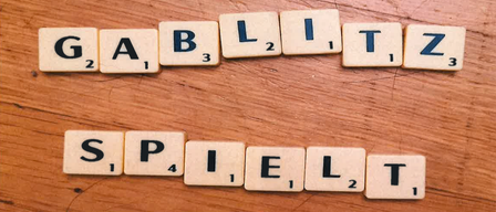 Eine hölzerne Oberfläche zeigt ein Scrabble-Spiel-Setup mit den Wörtern 'GABLIZ' und 'SPIELT', die aus Buchstabenfliesen gebildet werden. Jede Fliese hat eine Nummer, die ihren Punktewert angibt.
