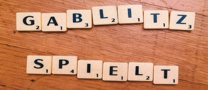 Eine hölzerne Oberfläche zeigt ein Scrabble-Spiel-Setup mit den Wörtern 'GABLIZ' und 'SPIELT', die aus Buchstabenfliesen gebildet werden. Jede Fliese hat eine Nummer, die ihren Punktewert angibt.