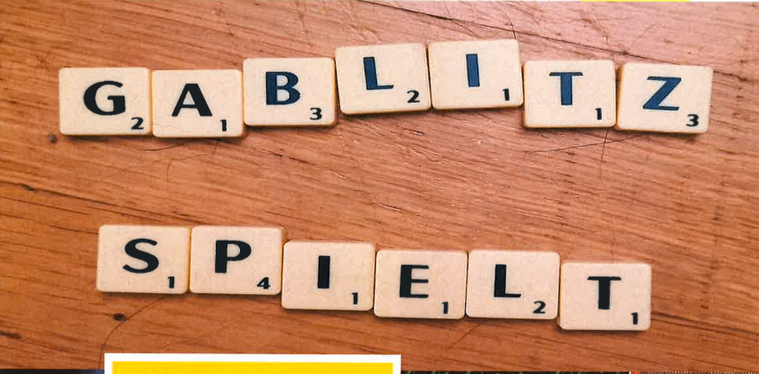 Eine hölzerne Oberfläche zeigt ein Scrabble-Spiel-Setup mit den Wörtern 'GABLIZ' und 'SPIELT', die aus Buchstabenfliesen gebildet werden. Jede Fliese hat eine Nummer, die ihren Punktewert angibt.