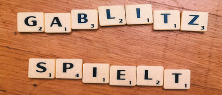 Scrabble tiles on a wooden surface spell out the words 'GABLIZT' and 'SPIELT'. The tiles have numbers and letters on them. The background is blurry.