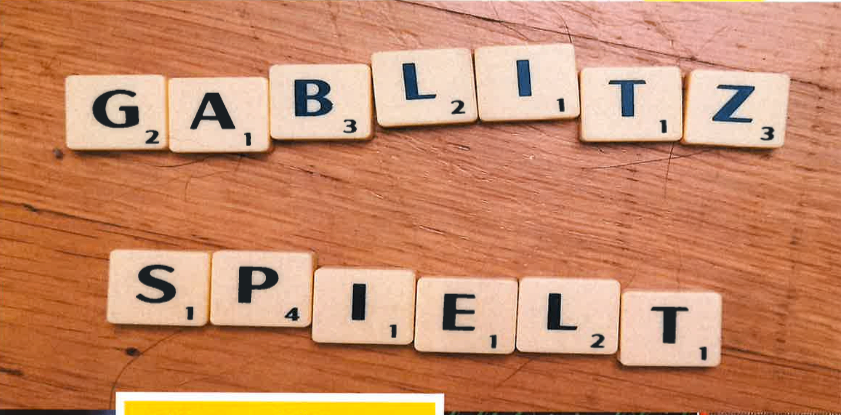 Scrabble-Steine auf einer Holzoberfläche bilden die Wörter 'GABLIZT' und 'SPIELT'. Die Steine haben Zahlen und Buchstaben darauf. Der Hintergrund ist unscharf.
