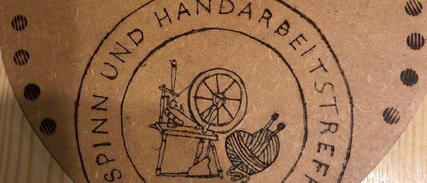 Eine Nahaufnahme eines braunen Kartons mit einem Logo. Das Logo zeigt eine Illustration einer Spinnmaschine mit Garn und einer Garnspule. Der Text lautet 'Spinn und Handarbeitstreff'.