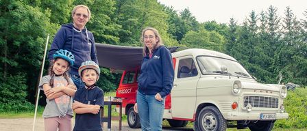 Eine vierköpfige Familie steht vor einem Oldtimer-Van. Die Kinder tragen Helme und Turnschuhe, die Erwachsenen Jacken und Brillen.