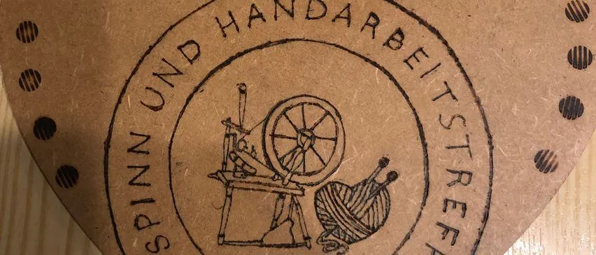 Ein braunes kreisförmiges Etikett mit einer vintage-stilisierten Illustration einer Spinnmaschine und Garn, umgeben von dem Text 'SPINNEN UND HANDARBEITSSTREIF'. Das Etikett hat eine leicht strukturierte Oberfläche.