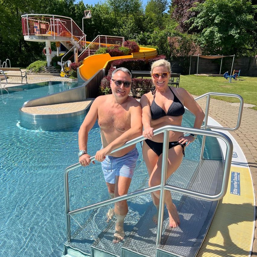 Ein Mann und eine Frau stehen lächelnd am Pooleingang. Hinter ihnen ist eine gelbe Wasserrutsche und ein Pool zu sehen.