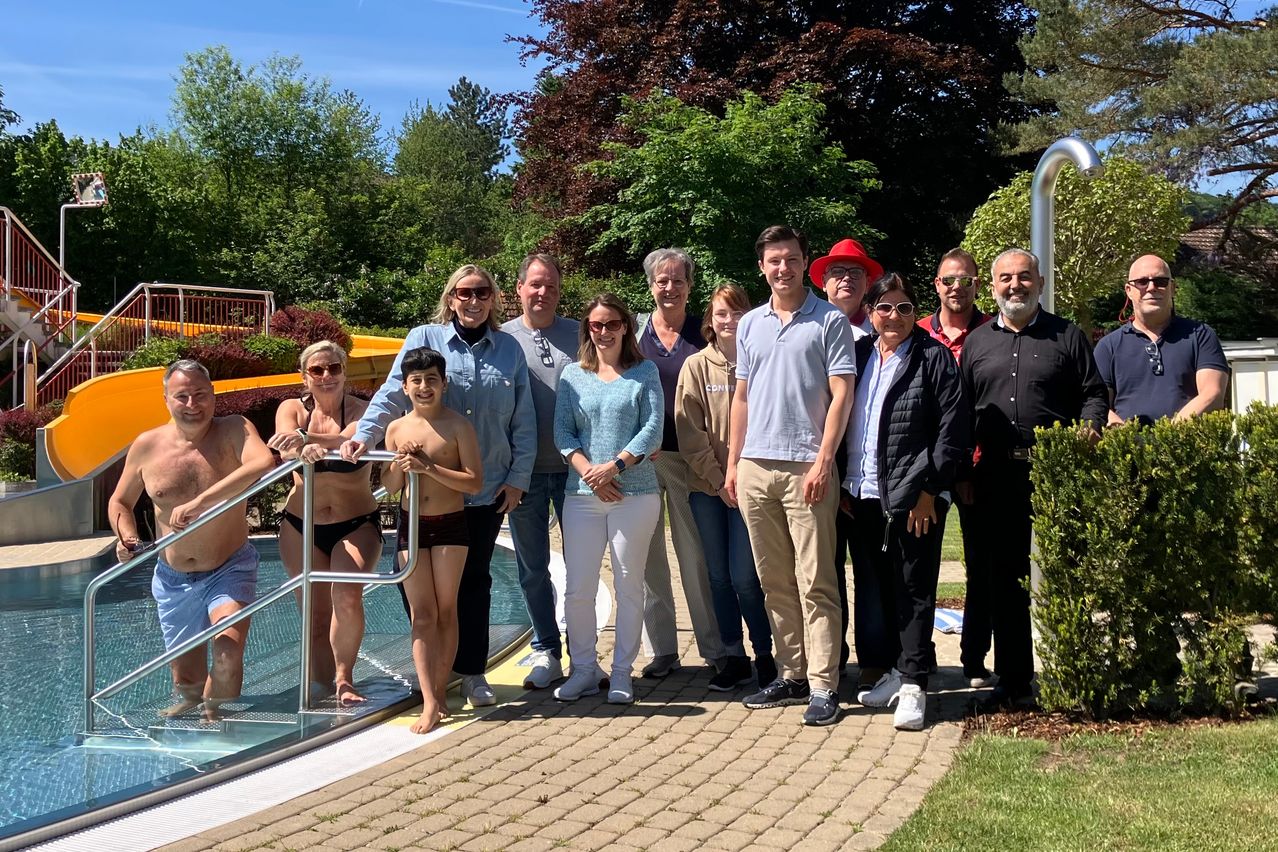 Eine Gruppe von Menschen in Badekleidung, einige im Pool und andere am Rand, posiert für ein Foto in einem Garten mit Bäumen und einer Rutsche im Hintergrund.