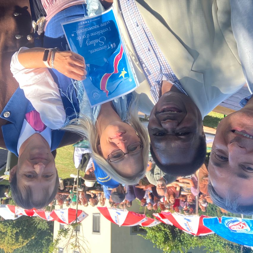 Vier Personen, darunter eine Frau mit einem blauen Büchlein, lächeln für die Kamera bei einer Outdoor-Veranstaltung mit Zelten und einer Menschenmenge im Hintergrund.
