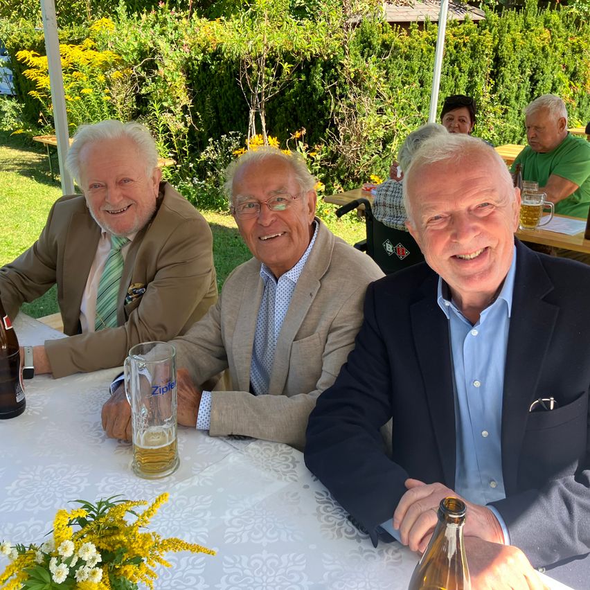 Drei ältere Männer sitzen lächelnd an einem Tisch. Sie befinden sich in einem Garten mit einer Hecke und einem Tisch mit einem Bierkrug. Einer von ihnen hält eine Flasche.