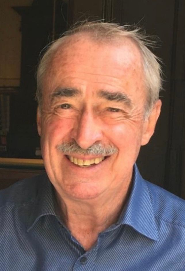 Ein älterer Mann mit weißem Haar und einem Schnurrbart lächelt. Er trägt ein blaues Hemd mit Kragen.