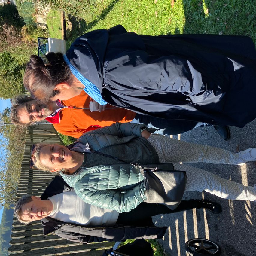 Fünf Menschen lächeln und stehen auf einem Weg. Hinter ihnen befindet sich ein Holzzaun. Links sind Bäume und Pflanzen. Ein Auto und ein Kinderwagen sind in der Nähe.