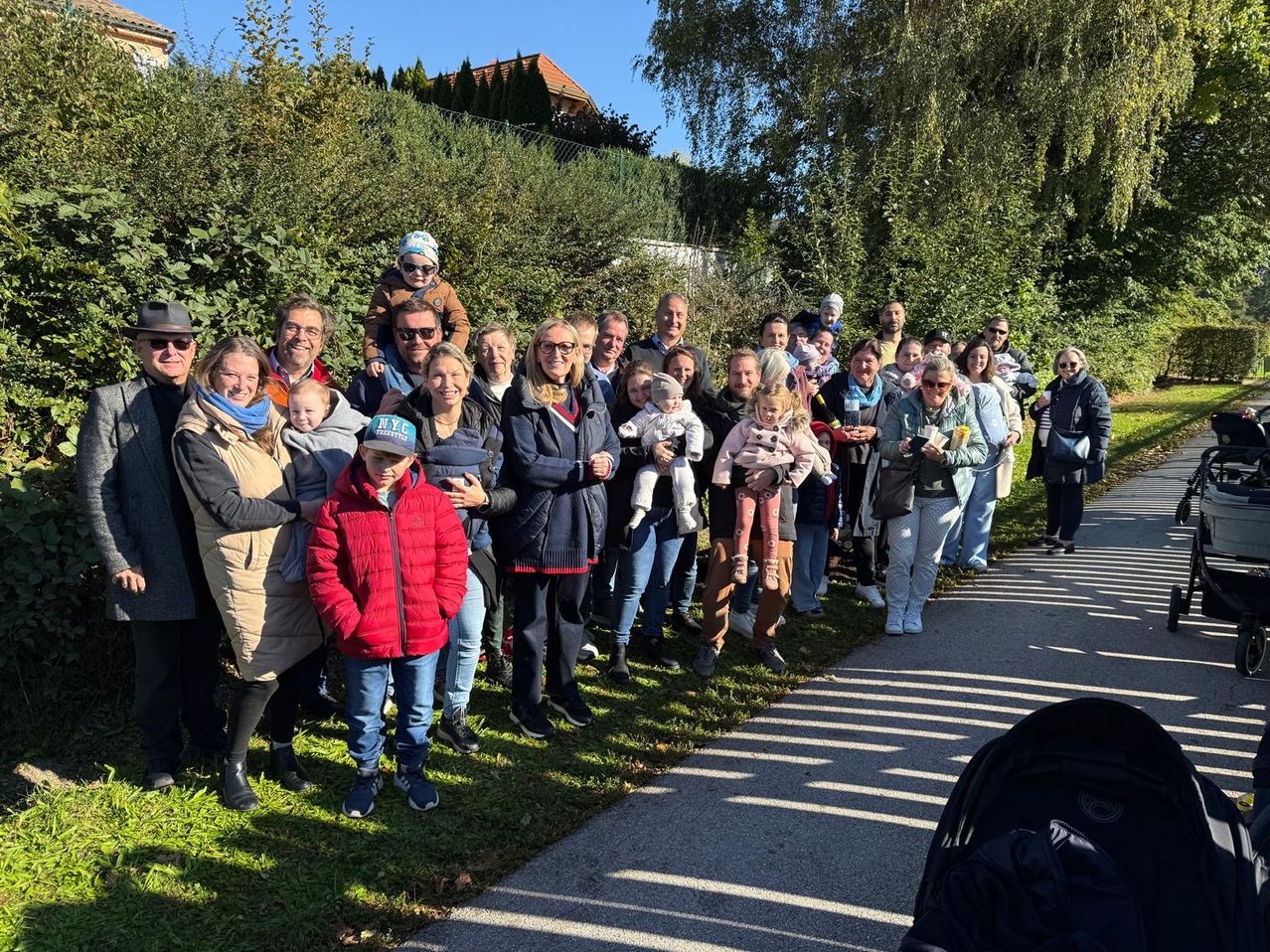 Eine große Gruppe von Erwachsenen und Kindern steht auf einem Pfad in einem Park, umgeben von Grün und blauem Himmel. Einige halten Babys.