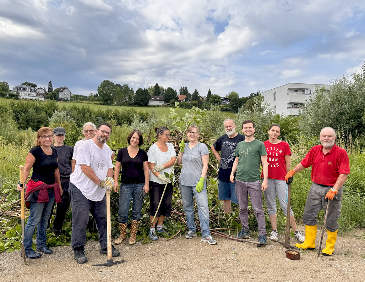 Eine Gruppe von Menschen steht draußen mit Gartenwerkzeugen und lächelt. Sie sind in der Nähe eines Busches, und einige tragen Handschuhe. Hinter ihnen ist ein Feld mit Bäumen und Häusern unter einem bewölkten Himmel sichtbar.