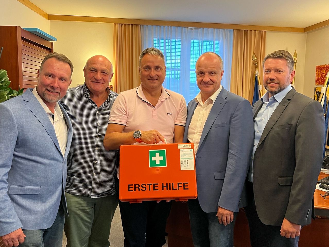Fünf Männer stehen in einer Innenumgebung. Der Mann in der Mitte hält eine rote Erste-Hilfe-Box. Der Mann auf der linken Seite trägt einen blauen Blazer und der Mann auf der rechten Seite trägt einen grauen Anzug.
