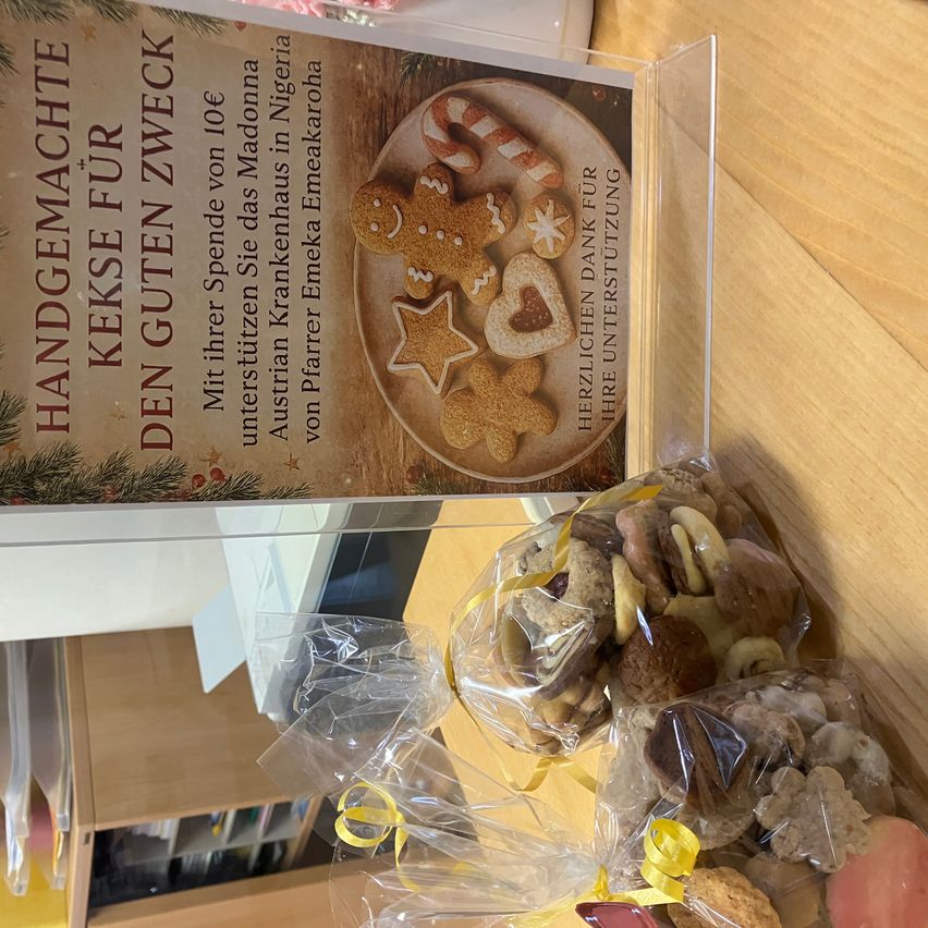 Ein durchsichtiges Plastikdisplay zeigt ein Weihnachtsbuch mit dem Titel 'Handgemachte Kekse für den guten Zweck' und einem Bild von dekorierten Keksen. Darunter befinden sich zwei Plastiktüten mit verschiedenen Keksen.