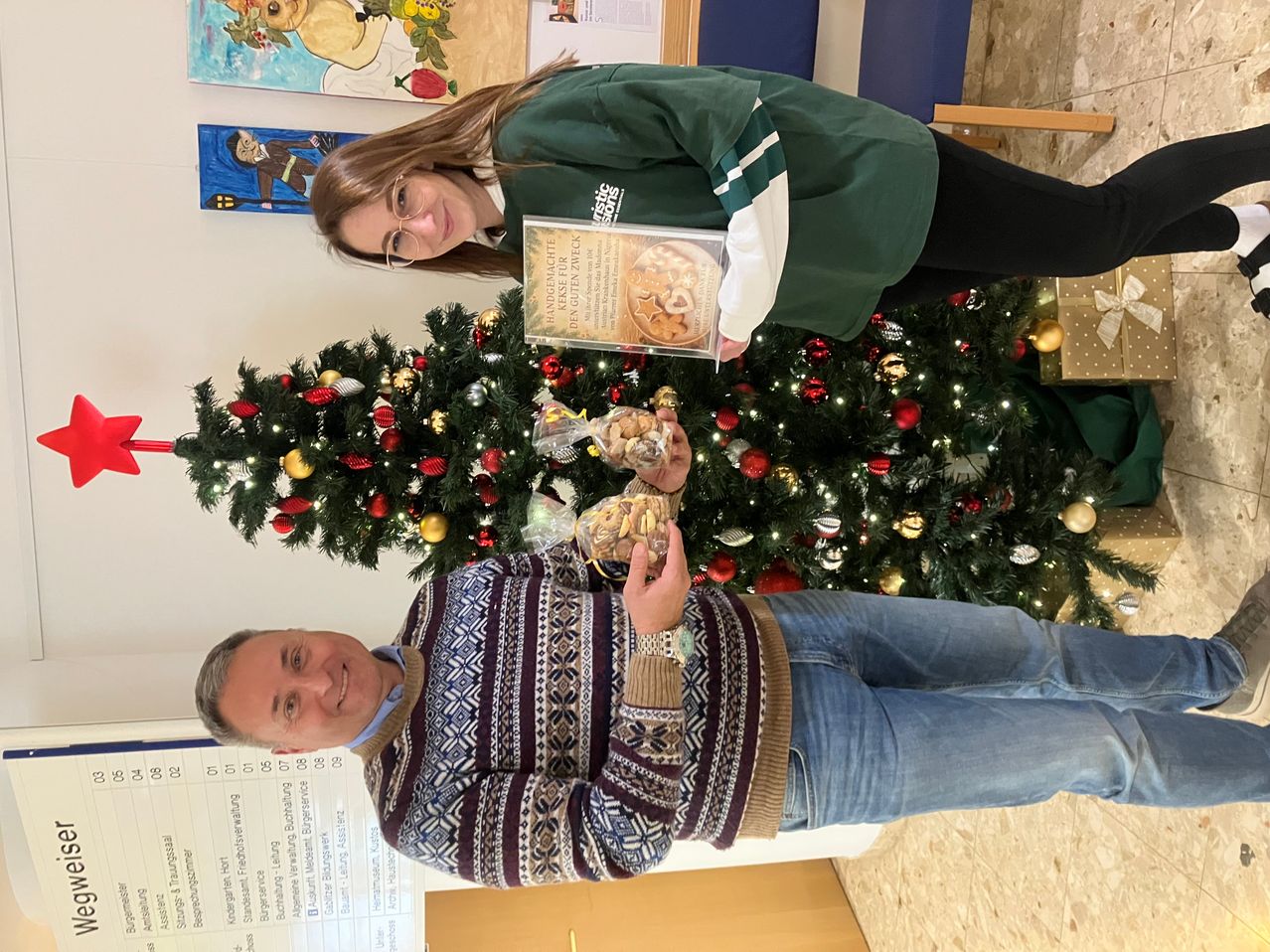 Ein Mann und eine Frau stehen vor einem geschmückten Weihnachtsbaum. Die Frau hält ein Schild und eine Tüte mit Keksen, während der Mann lächelt und eine Tüte mit Keksen in der Hand hält. Hinter ihnen sind zwei Gemälde an der Wand zu sehen.