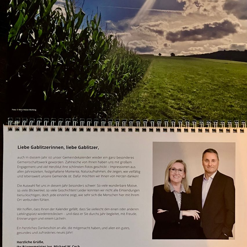 Ein Gemeindekalender mit einem Bild von einem Maisfeld und zwei Personen, von denen eine ein Mann und die andere eine Frau ist, beide mit Brille. Sie stehen eng beieinander und lächeln. Der Kalender hat eine Botschaft auf Deutsch.