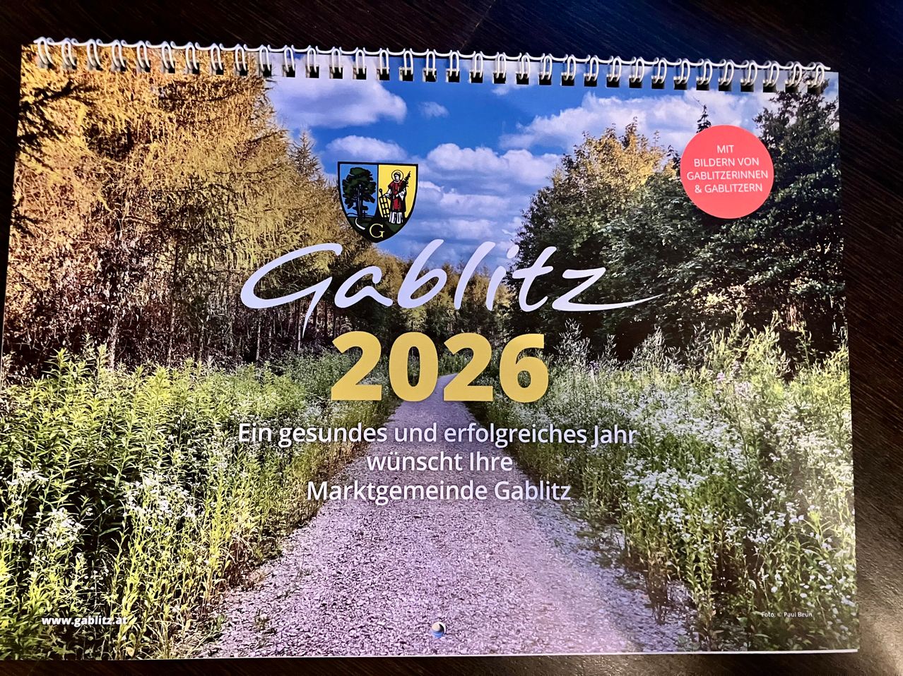 Ein 2026 Kalender aus Gablitz mit Bildern von Einheimischen und Bewohnern. Die Titelseite zeigt einen Feldweg mit Bäumen und Grün. Es steht 'Ein gesundes und erfolgreiches Jahr wünscht Ihre Marktgemeinde Gablitz.'