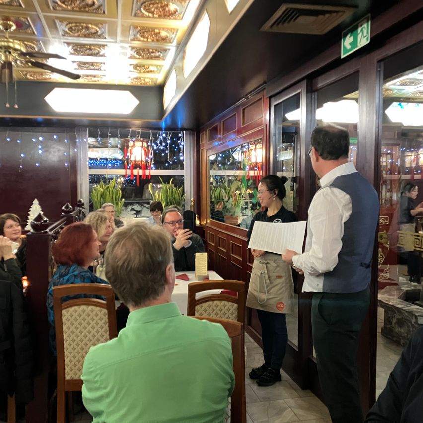 Das Innere eines Restaurants mit sitzenden Gästen. Eine Frau hält ein Menü, und ein Mann in einer Weste spricht mit ihr. Der Raum ist mit Weihnachtsthemen dekoriert.