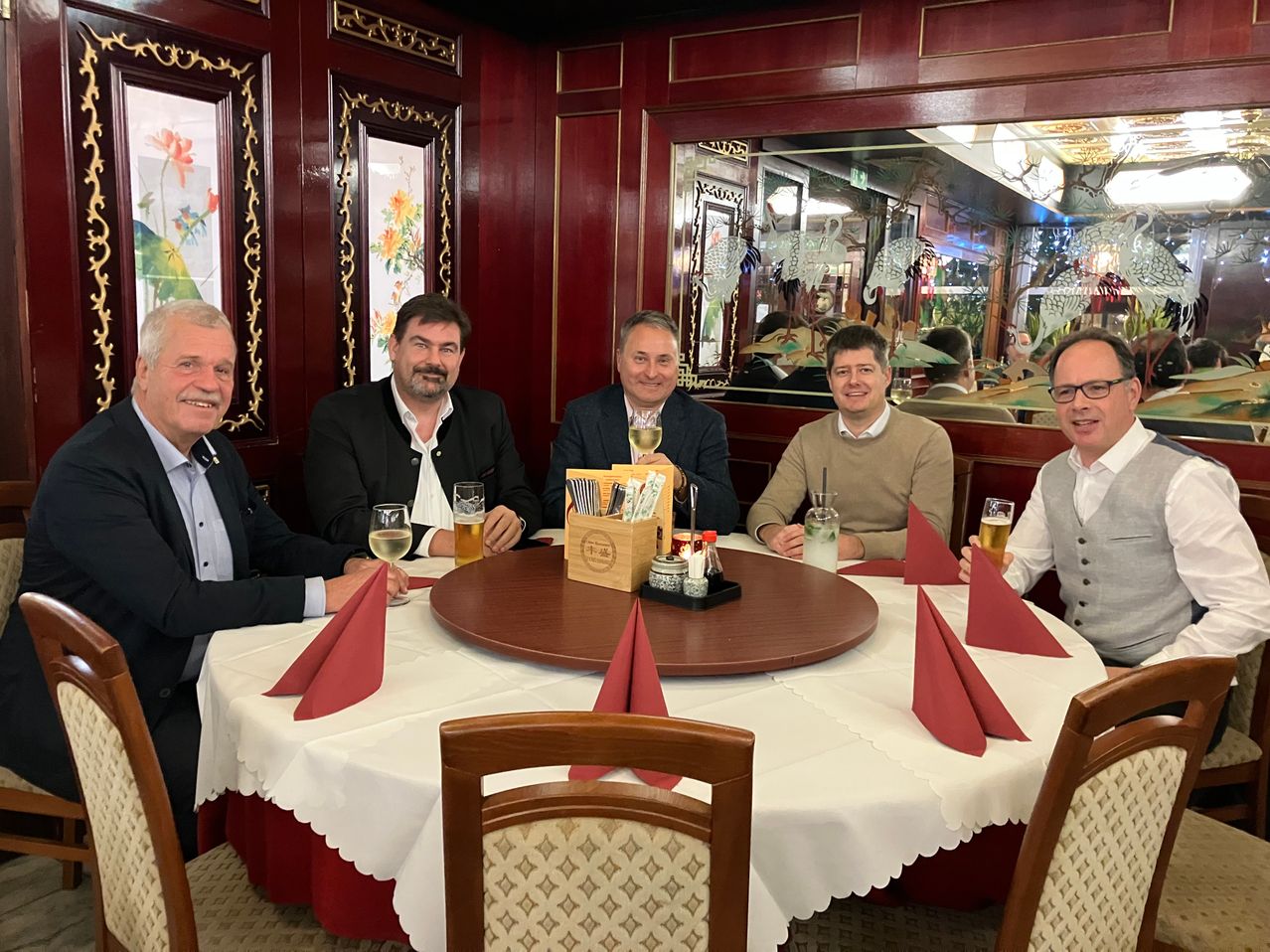 Fünf Männer sitzen um einen Tisch herum, lächeln, mit Getränken und Essen vor sich. Der Tisch ist mit roten Servietten, Weingläsern und einer Holzbox gedeckt. Im Hintergrund befindet sich ein Spiegel mit Reflexionen von Menschen und einer Kristallleuchte.