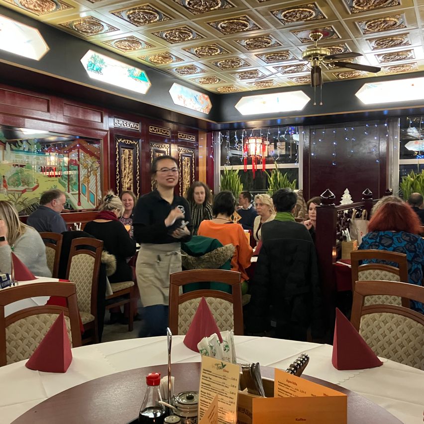 Ein Restaurant mit einer Frau, die an einem Tisch steht. Der Raum hat Holzstühle und Tische mit Servietten. Die Decke hat einen Ventilator und dekorative Lichter.