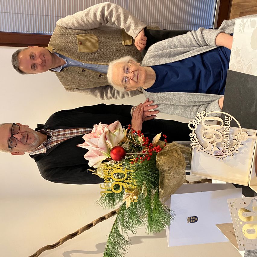 Zwei Menschen stehen eng beieinander mit einem Blumenstrauß und Geburtstagskarten vor ihnen. Ein Mann trägt eine Brille und eine Krawatte.