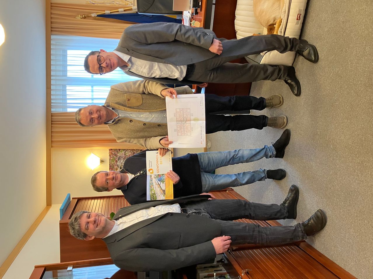 Vier Männer in formeller Kleidung stehen in einem Raum. Einer hält ein Zertifikat, ein anderer ein Dokument. Sie lächeln und einer lehnt sich gegen eine Wand.