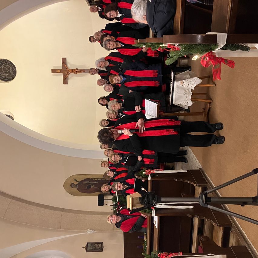 Ein Chor in roten und schwarzen Gewändern, der in einer Kirche steht, einige halten Musikblätter, ein Kreuz an der Wand und eine Weihnachtsdekoration.