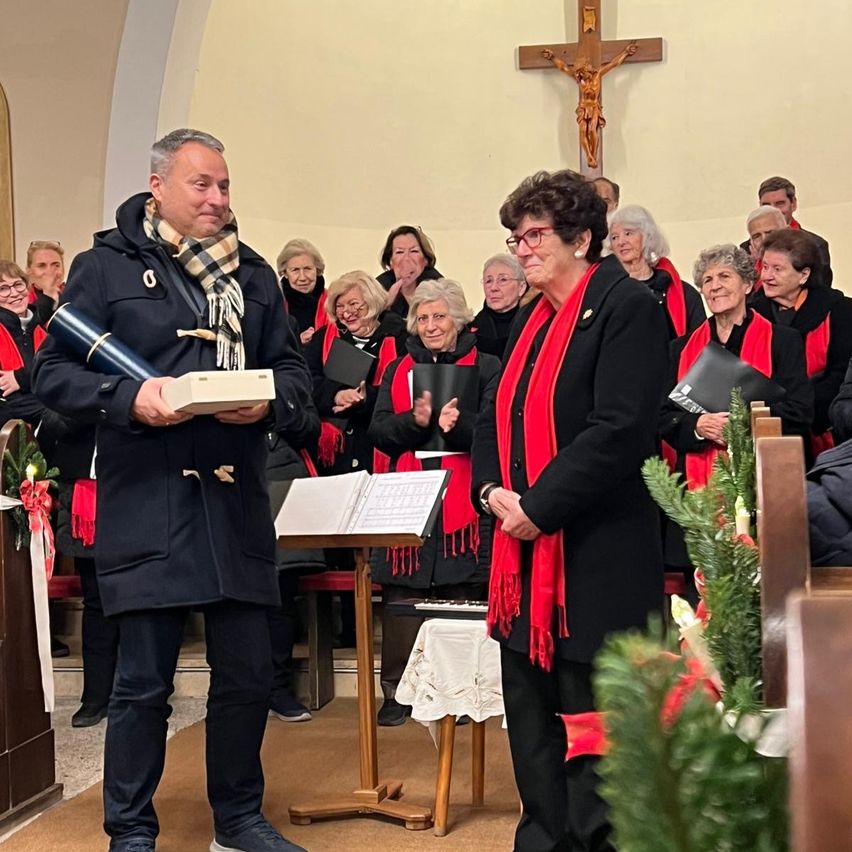 Ein Chor in roten Schals steht in einer Kirche, ein Mann in einem Schal hält ein Geschenk. Eine Frau mit Brille und roter Schürze steht in der Nähe, und ein Kreuz ist an der Wand.