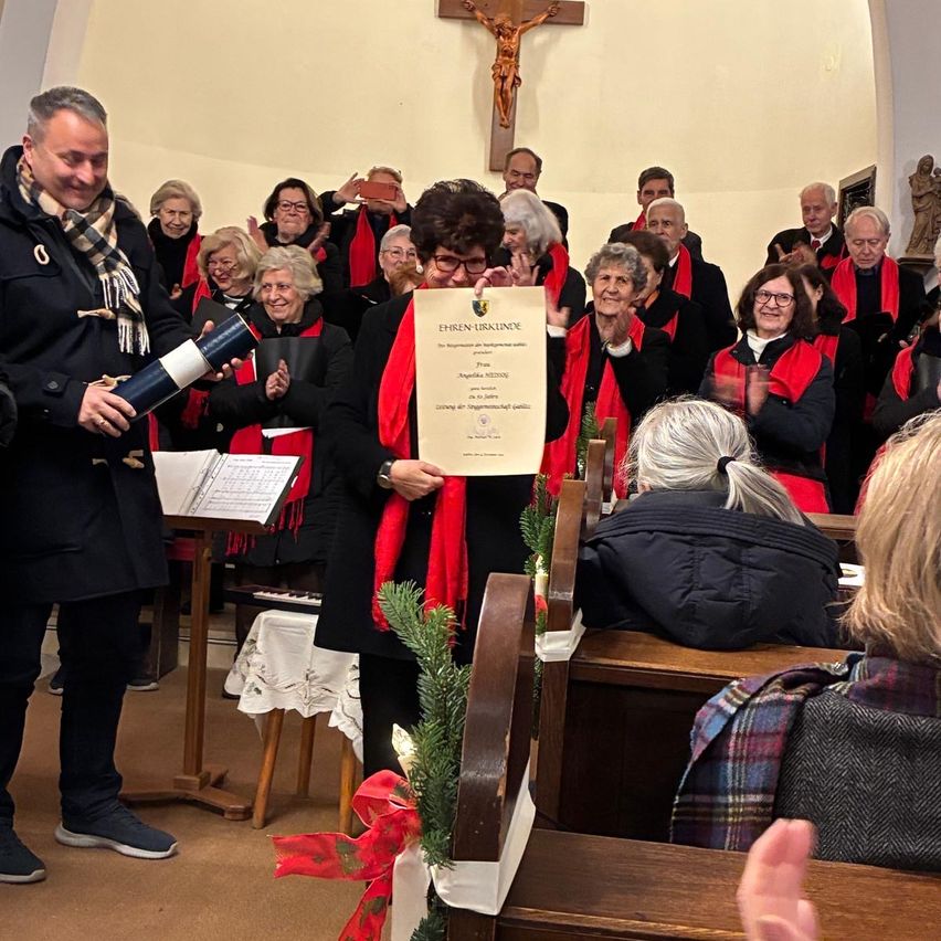 Ein Chor steht in einer Kirche und hält ein Zertifikat. Ein Mann überreicht es einer Frau vor ihnen.