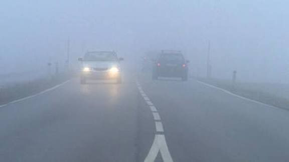 Zwei Autos fahren auf einer nebligen Straße mit eingeschalteten Scheinwerfern. Die Straße hat weiße Markierungen und einen weißen Pfeil, der nach vorne zeigt.