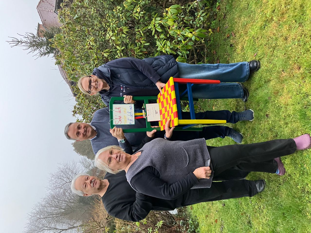 Vier Personen lächeln und posieren für ein Foto auf einem Rasen. Sie halten einen bunten Stuhl. Hinter ihnen sind Pflanzen und Bäume.