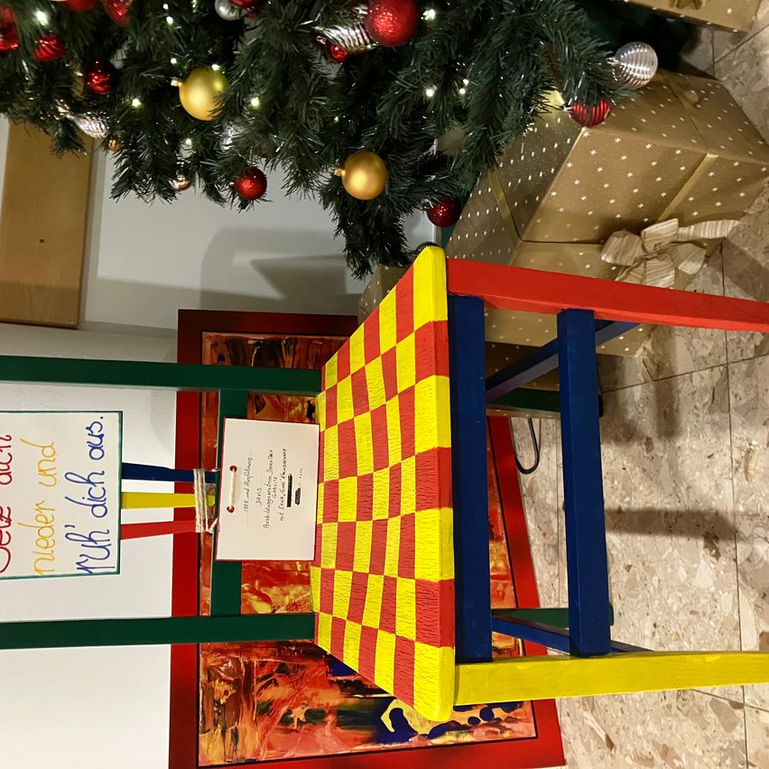 Ein bunter Stuhl steht vor einem Weihnachtsbaum auf dem Boden. Der Stuhl hat ein kariertes Muster. Ein weißes Papier ist mit einem Band am Stuhl befestigt. Hinter dem Stuhl befindet sich ein in Goldpapier eingewickeltes Geschenk.