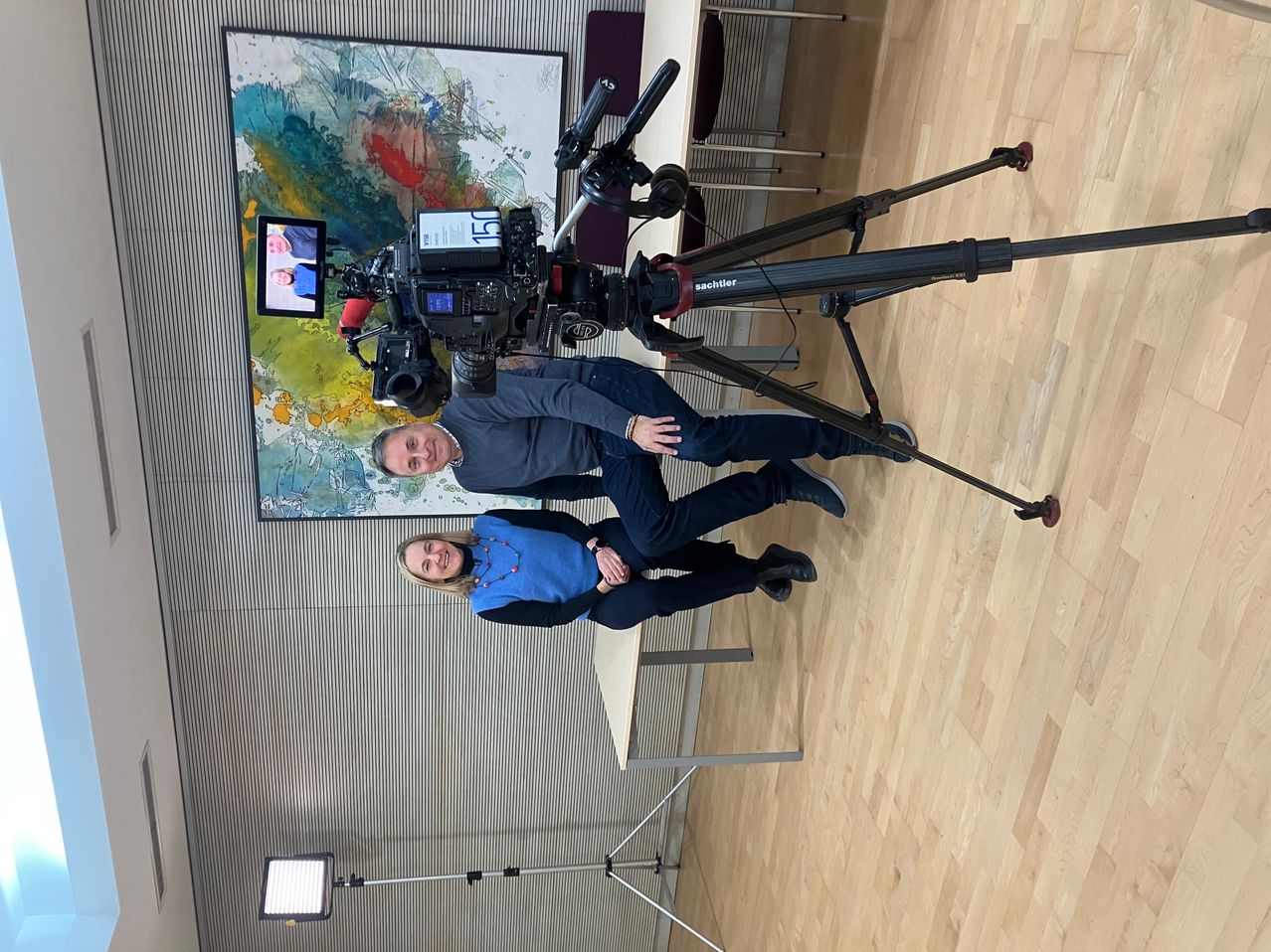 Zwei Personen sitzen unter einer Kamera auf einem Stativ und lächeln in die Kamera. Hinter ihnen ziert ein farbenfrohes Gemälde die Wand.