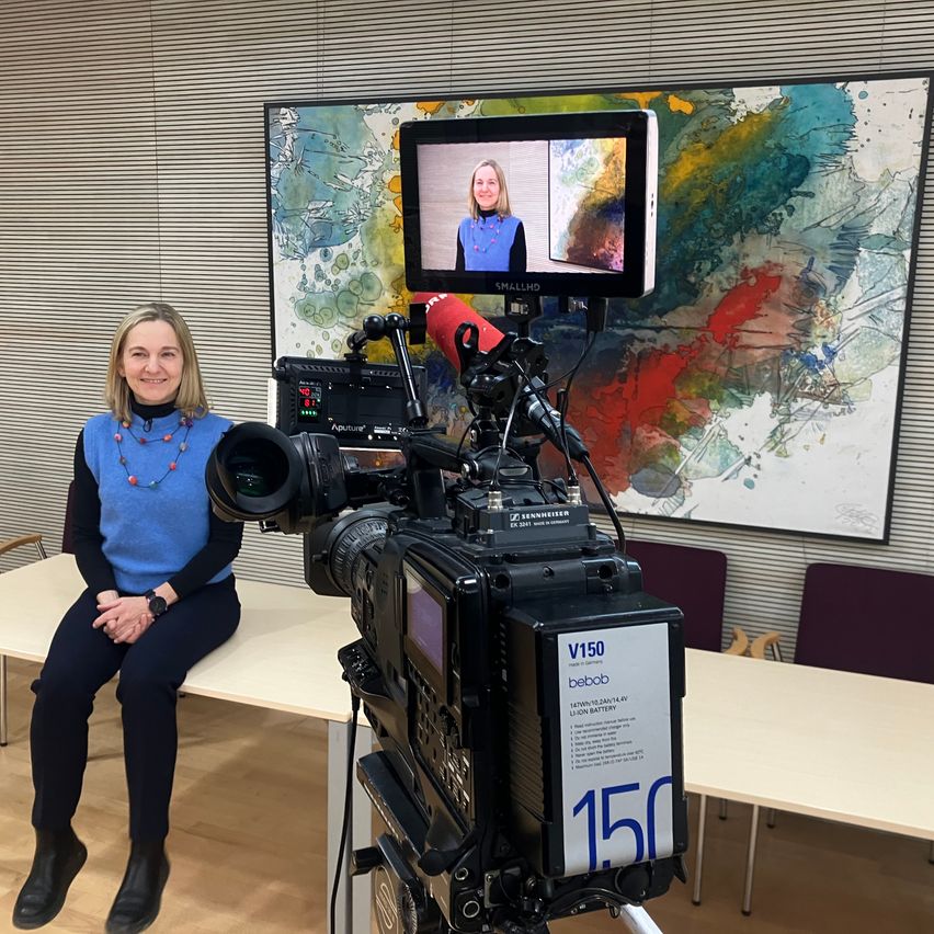 Eine Frau sitzt auf einer Bank vor einer Kamera-Einrichtung. Hinter ihr ist ein großes, farbenfrohes Gemälde an der Wand. Die Kamera ist auf einem Stativ montiert, mit einem Monitor, der eine Frau anzeigt.