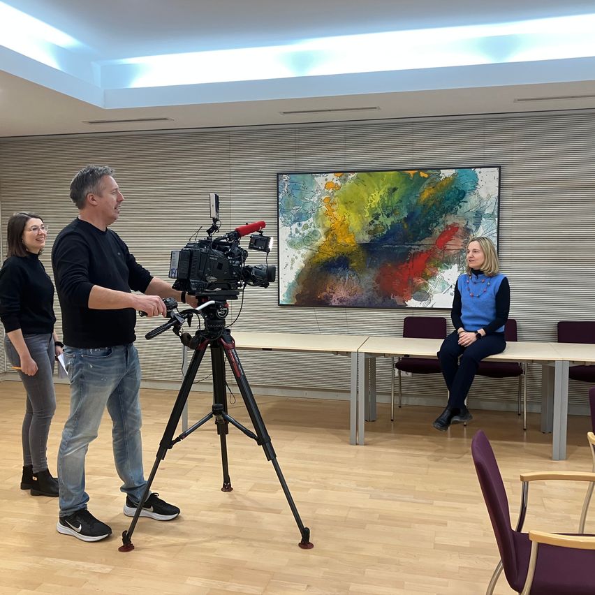 Ein Mann, der eine Kamera hält, steht in einem Raum und filmt eine Frau, die an einem Schreibtisch sitzt. Eine weitere Frau steht neben dem Kameramann, und es gibt ein großes abstraktes Gemälde an der Wand.